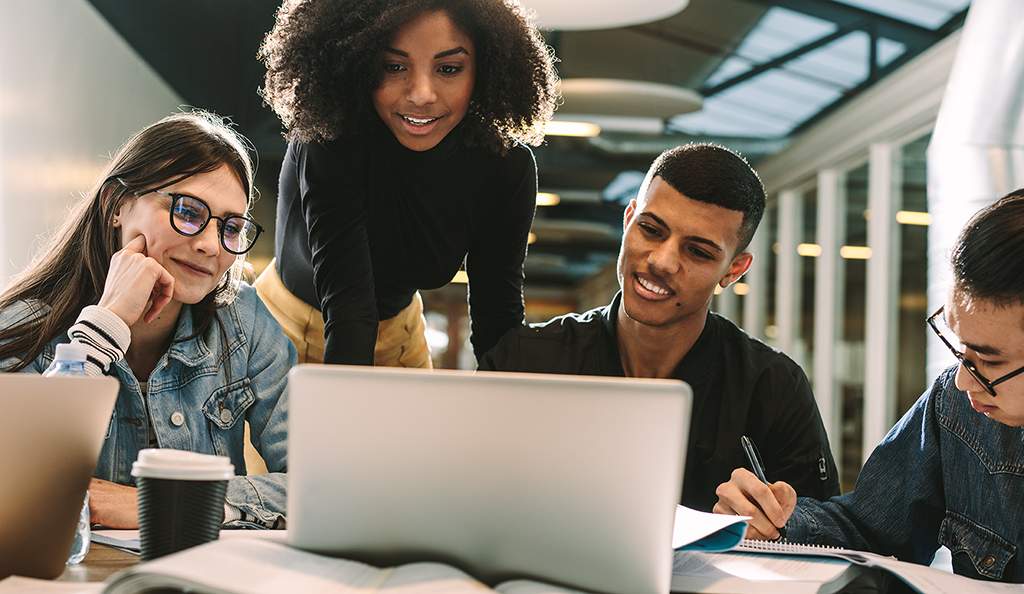 Três estudantes olham para um laptop, enquanto um quarto estudante faz anotações