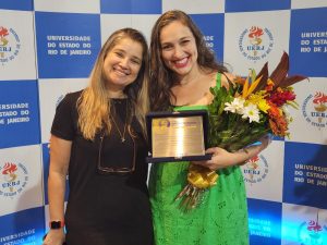 A imagem mostra as servidoras Marcia Taborda e Débora Milagres, sorrindo. Débora está segurando uma placa de premiação e um ramo de flores.